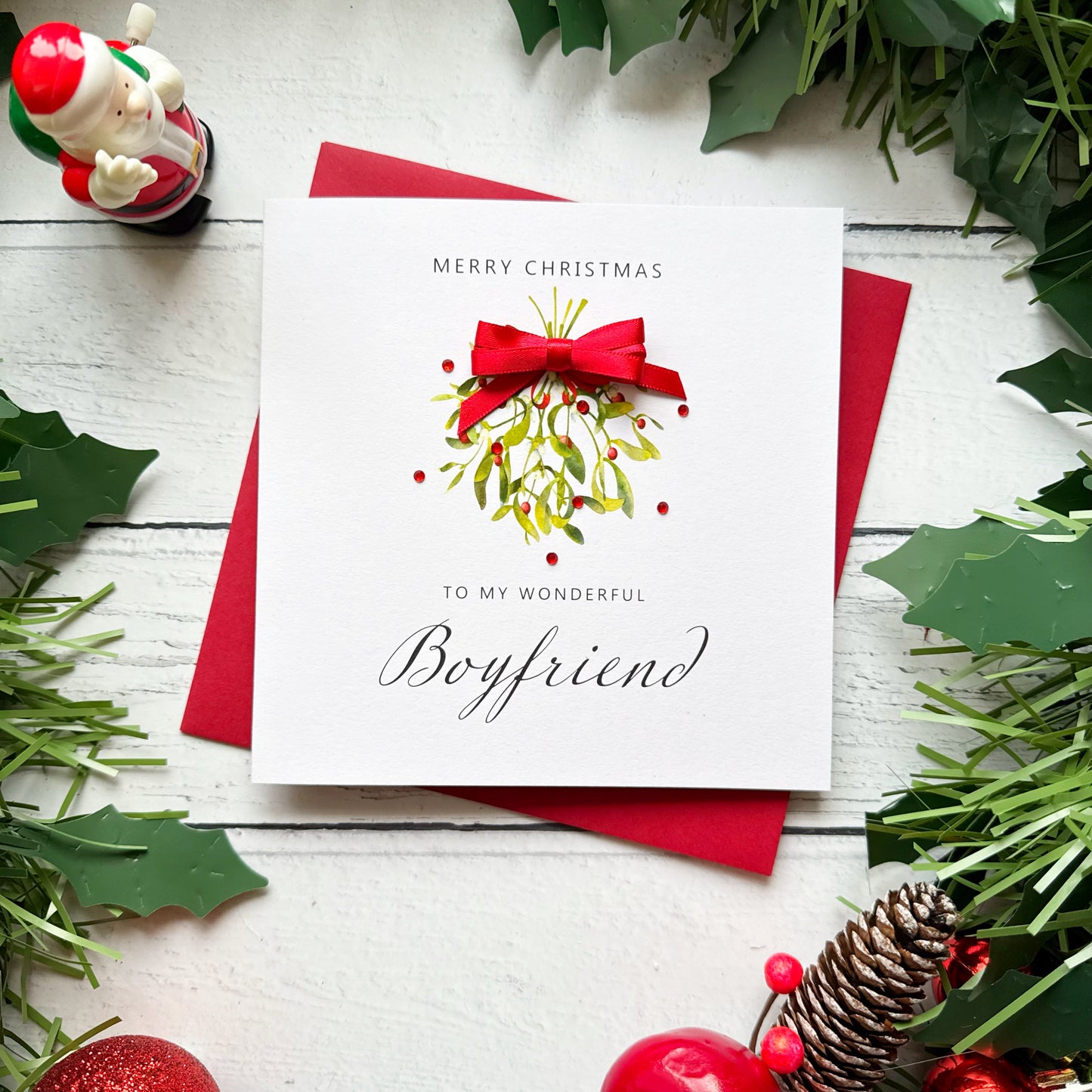 A square white Christmas greeting card with a red bow and mistletoe illustration, reading 'Merry Christmas to my wonderful Boyfriend,' placed on a red envelope on a white wood surface with holly and holiday decorations around it.