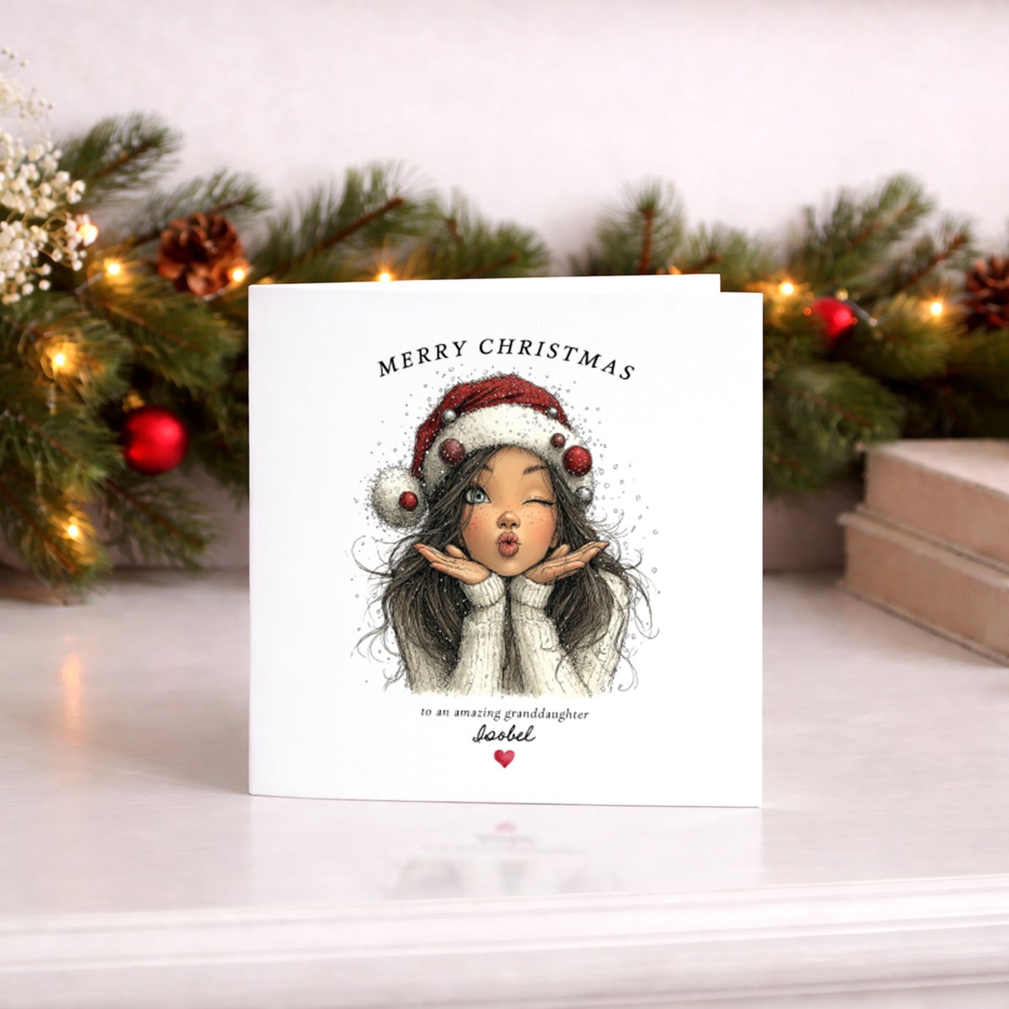 A square white Christmas card featuring an illustration of a dark-haired girl in a red Santa hat decorated with red and silver baubles, wearing a cream knit jumper and blowing a kiss with glitter and snowflakes around her. Text reads "Merry Christmas to an amazing granddaughter" with "Isobel" in cursive script and a small red heart. Displayed against a lit pine garland with red baubles.