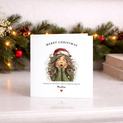 A personalized Christmas greeting card with an illustration of a surprised girl in a green sweater and Santa hat, standing on a white surface with a blurred festive background of pine branches and warm lights.