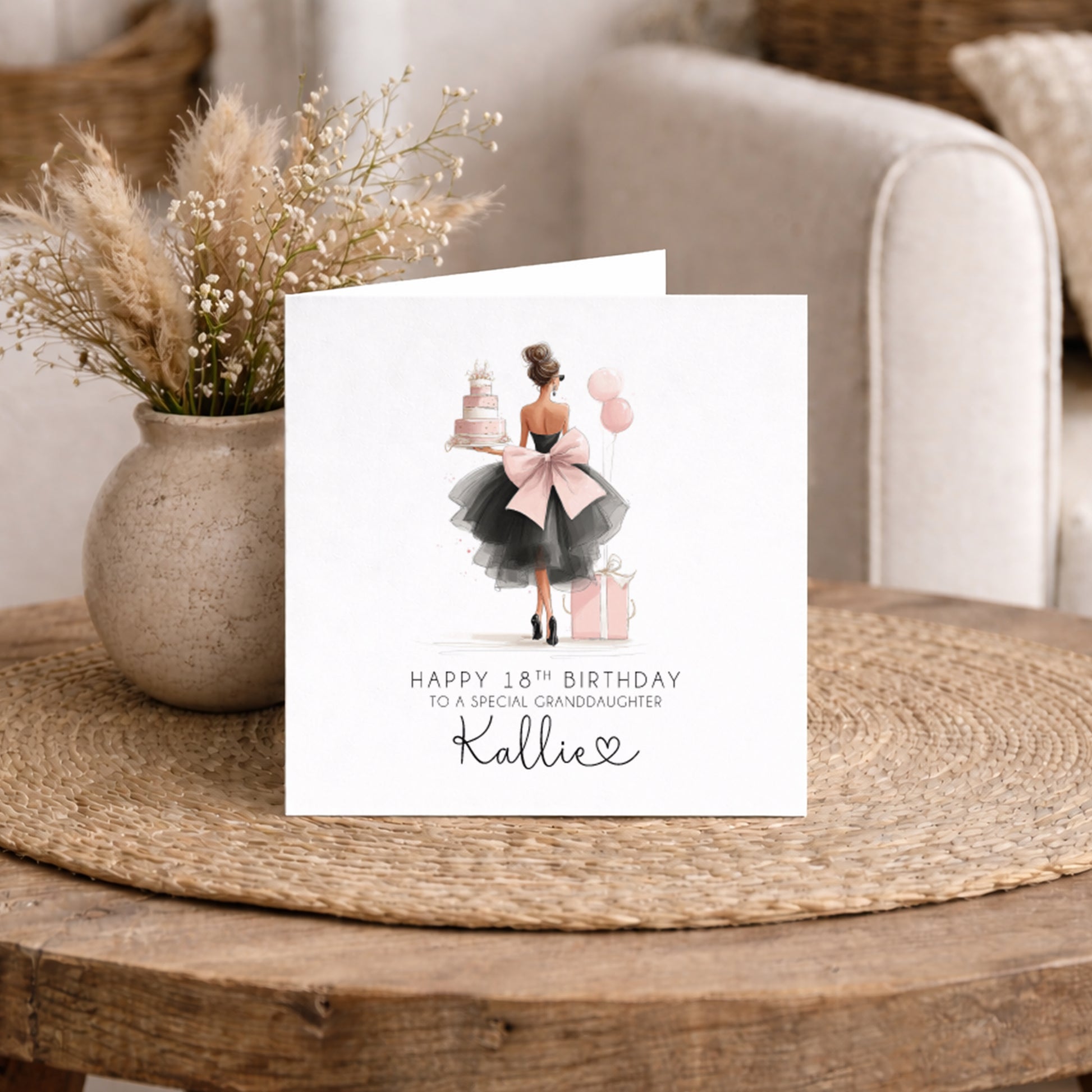 A square white 18th birthday card featuring an illustration of a young woman viewed from behind in a black tulle dress with a large pink bow, carrying a tiered pink birthday cake, with pink balloons and a gift box beside her. Text reads "Happy 18th Birthday to a Special Granddaughter" with "Kallie" in cursive script and a small heart. Displayed on a wicker table with pampas grass and dried flowers.
