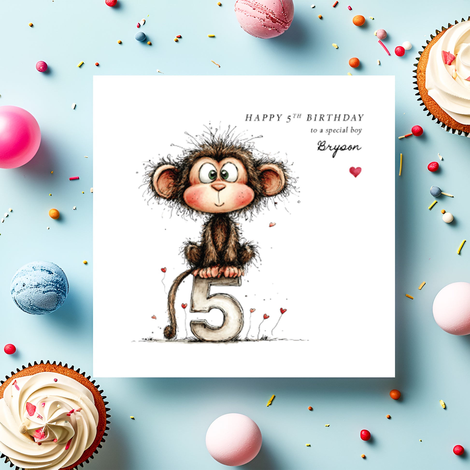 A square white birthday card featuring an illustration of a cheeky wide-eyed monkey with rosy cheeks and scruffy fur, perched on a large number five with small red heart details scattered around. Text reads "Happy 5th Birthday to a special boy" with "Bryson" in cursive script and a small red heart. Displayed on a light blue surface with cupcakes, macarons and colourful sprinkles.