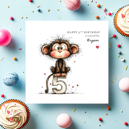 A square white birthday card featuring an illustration of a cheeky wide-eyed monkey with rosy cheeks and scruffy fur, perched on a large number five with small red heart details scattered around. Text reads "Happy 5th Birthday to a special boy" with "Bryson" in cursive script and a small red heart. Displayed on a light blue surface with cupcakes, macarons and colourful sprinkles.