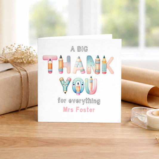 Personalised thank you teacher greeting card featuring colourful stationery-themed lettering spelling “Thank You” with the custom name “Mrs Foster.” The card reads “A Big Thank You for everything” and is displayed on a wooden table beside wrapped gifts and stationery items.
