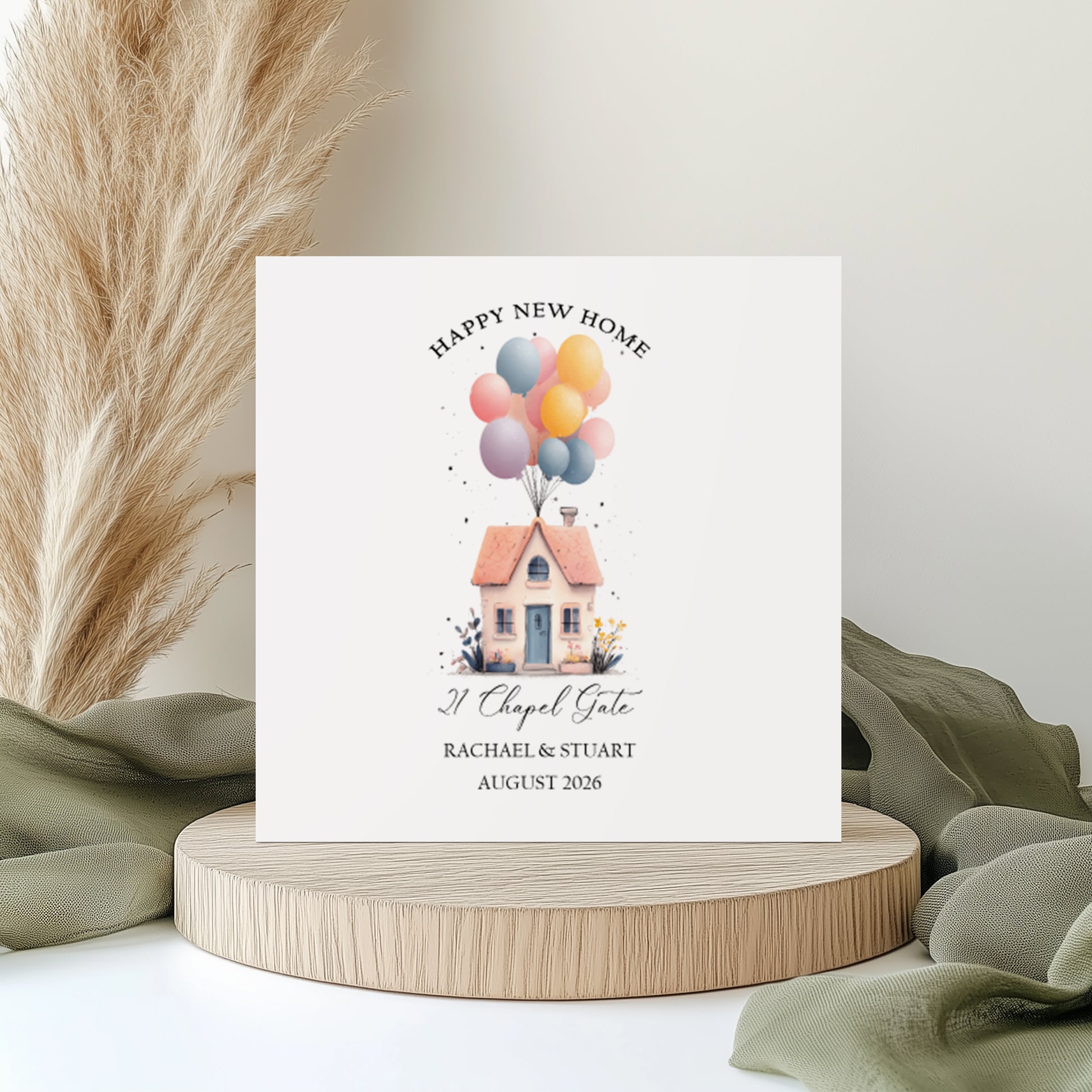 A square white new home card featuring a watercolour illustration of a pretty cottage with a terracotta roof, blue front door and garden flowers, lifted by a large bunch of pastel balloons in pink, yellow, blue and lilac. Text reads "Happy New Home" in arched capitals, with "21 Chapel Gate" in cursive script, "Rachael & Stuart" and "August 2026" below. Displayed on a wooden disc with sage green linen and pampas grass.