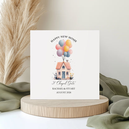 A square white new home card featuring a watercolour illustration of a pretty cottage with a terracotta roof, blue front door and garden flowers, lifted by a large bunch of pastel balloons in pink, yellow, blue and lilac. Text reads "Happy New Home" in arched capitals, with "21 Chapel Gate" in cursive script, "Rachael & Stuart" and "August 2026" below. Displayed on a wooden disc with sage green linen and pampas grass.