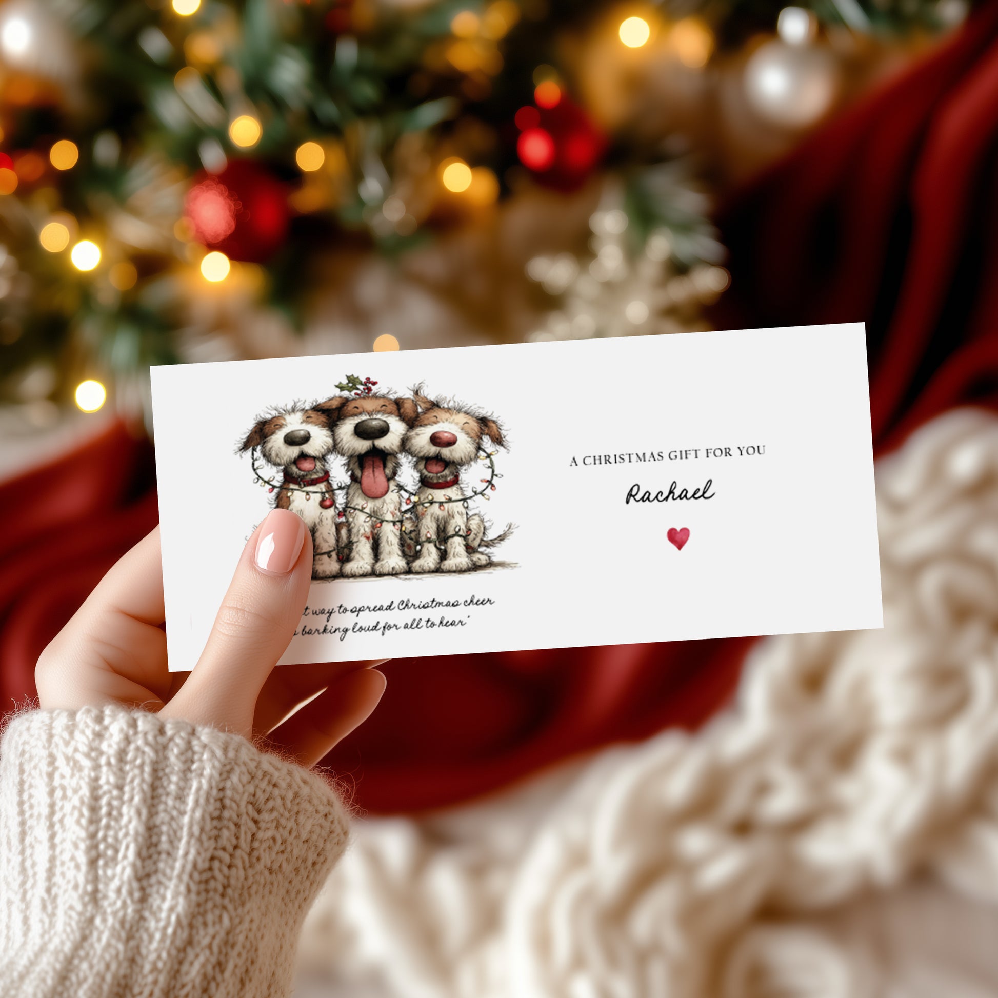 A rectangular white Christmas money wallet held in hand, featuring three scruffy dogs tangled in colourful fairy lights with a holly sprig on top and the sentiment about spreading Christmas cheer. To the right, "A Christmas Gift for You" in spaced capitals with "Rachael" in cursive script and a small red heart. Displayed against a blurred Christmas tree with red and gold baubles.