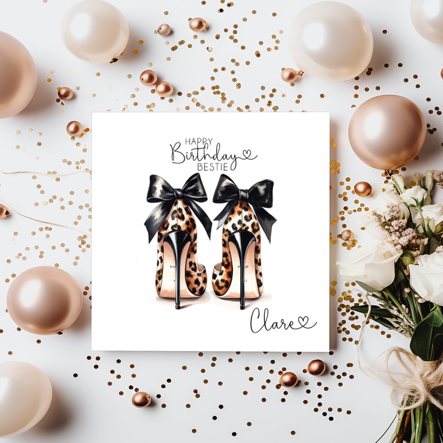 A square white birthday card featuring an illustration of two leopard print high heeled shoes, viewed from behind, each decorated with a black satin bow. The text "Happy Birthday Bestie" is printed in black script and capitals at the top of the card, with a small heart, and the name "Clare" written in cursive script in the bottom corner alongside another small heart. The card is displayed on a white surface surrounded by rose gold and cream balloons, scattered gold confetti and pearl beads, with white roses
