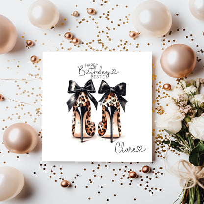 A square white birthday card featuring an illustration of two leopard print high heeled shoes, viewed from behind, each decorated with a black satin bow. The text "Happy Birthday Bestie" is printed in black script and capitals at the top of the card, with a small heart, and the name "Clare" written in cursive script in the bottom corner alongside another small heart. The card is displayed on a white surface surrounded by rose gold and cream balloons, scattered gold confetti and pearl beads, with white roses