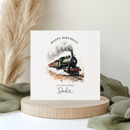 Square birthday card featuring a vintage steam train illustration with smoke billowing from the engine. The card reads “Happy Birthday to a train-mendous Dad” in elegant typography. Displayed on a neutral styled background with pampas grass and soft green fabric accents.