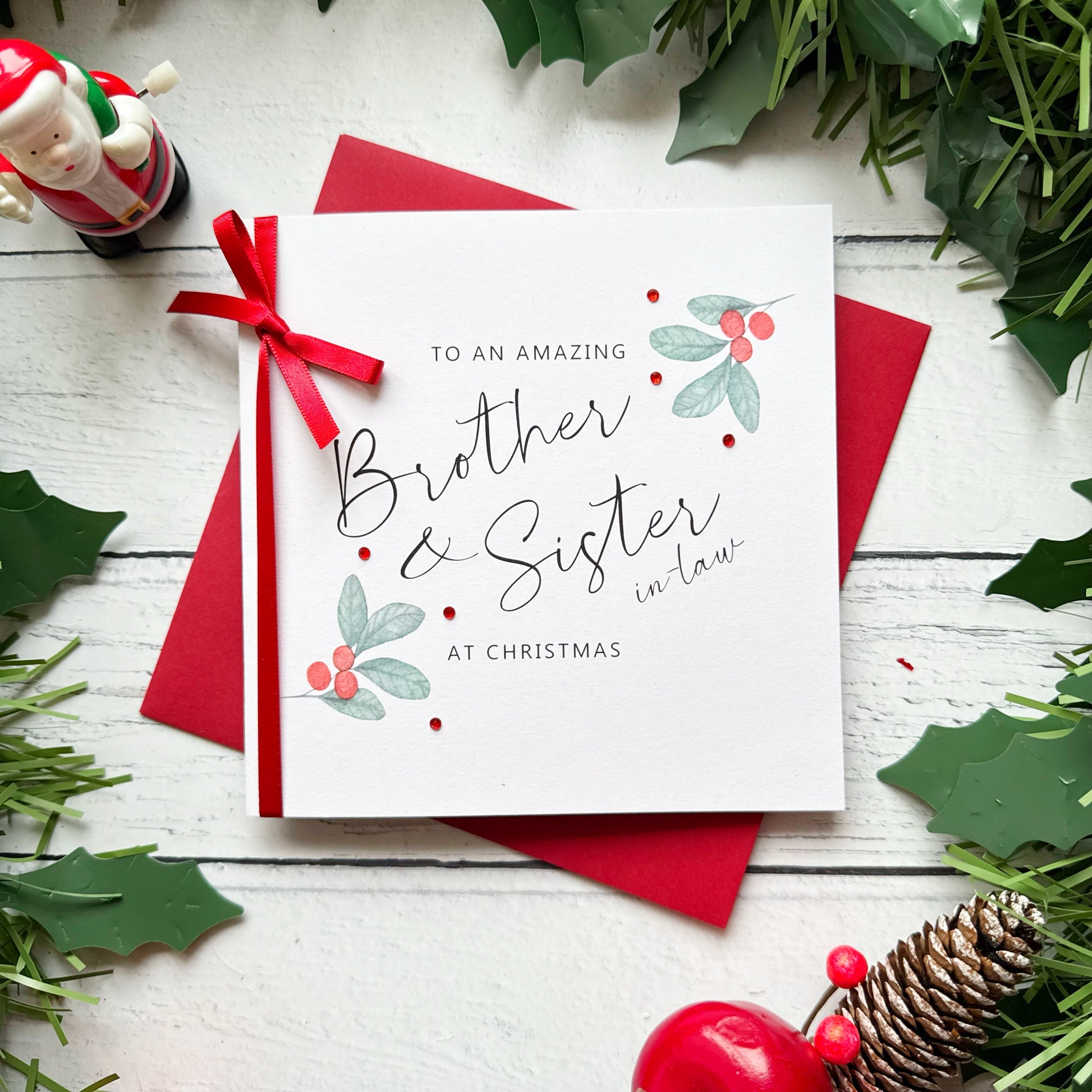 A square white Christmas card featuring watercolour holly berry sprigs with sage green leaves and red berries in the upper right and lower left corners, accented with small scattered red gem embellishments. The text reads "to an Amazing" in small spaced capitals at the top, with "Brother & Sister-in-Law" in large flowing black cursive script in the centre, and "at Christmas" in small spaced capitals below. The card is tied with a real red satin ribbon bow on the left side and sits on a matching red envelope