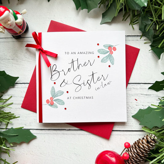 A square white Christmas card featuring watercolour holly berry sprigs with sage green leaves and red berries in the upper right and lower left corners, accented with small scattered red gem embellishments. The text reads "to an Amazing" in small spaced capitals at the top, with "Brother & Sister-in-Law" in large flowing black cursive script in the centre, and "at Christmas" in small spaced capitals below. The card is tied with a real red satin ribbon bow on the left side and sits on a matching red envelope