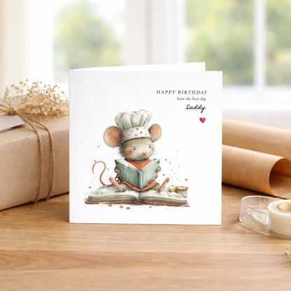 A square white birthday card featuring an illustration of a small mouse wearing a chef’s hat and orange apron, sitting on an open recipe book dusted with flour, reading intently with tiny stars scattered around. Text reads “Happy Birthday have the best day” in spaced capitals and small italics, with “Daddy” in flowing cursive script and a small red heart. Displayed on a wooden surface with kraft wrapping and dried flowers.