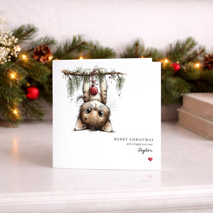 A white Christmas greeting card featuring a whimsical illustration of a cat hanging upside down from a branch with ornaments, with the text 'MERRY CHRISTMAS and a happy new year' and the name 'Taylor' printed below. The card is displayed on a light surface with a blurred Christmas garland in the background.