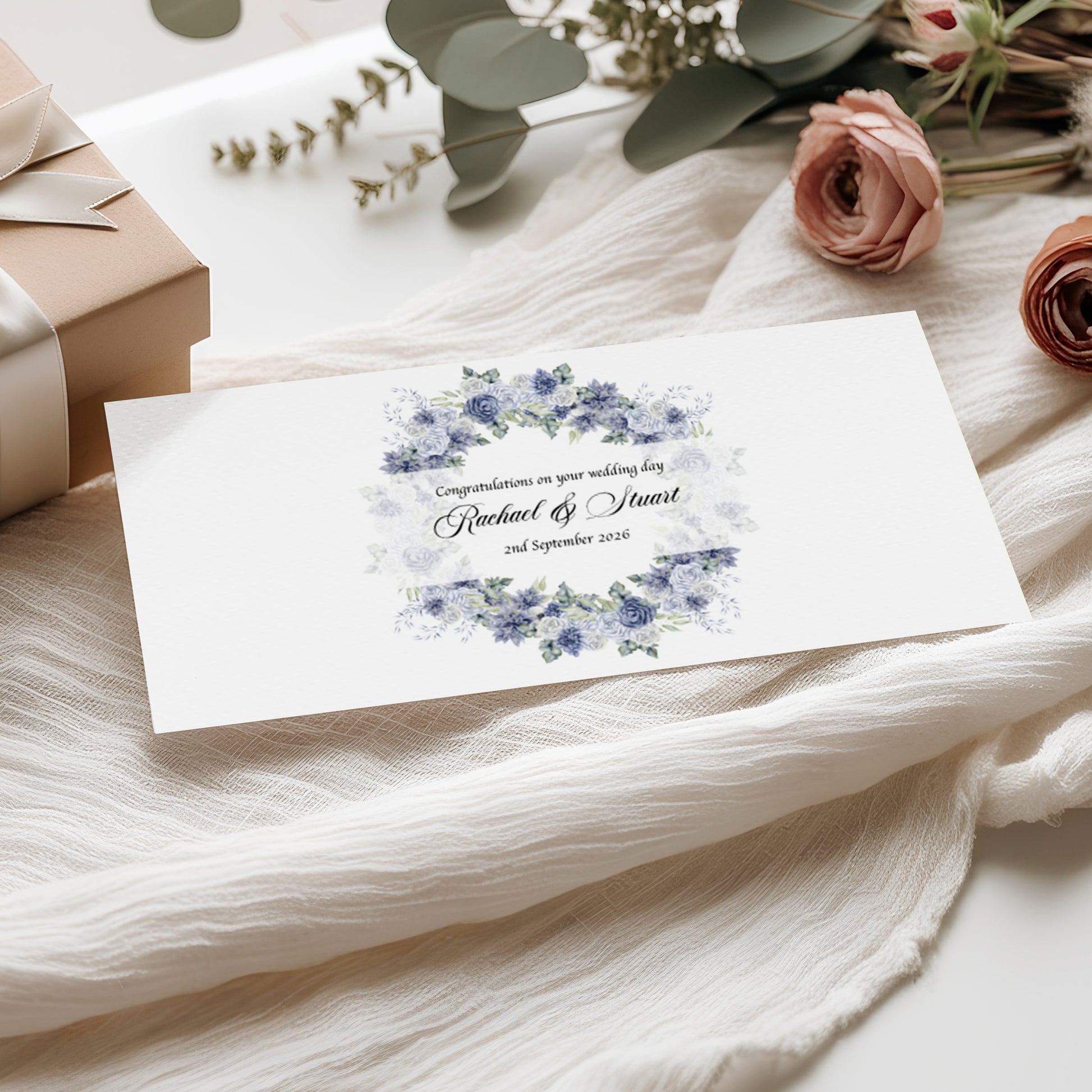Wedding money wallet card with blue floral design with names and wedding date on a white fabric background with flowers and gift box.