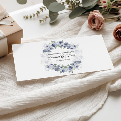Wedding money wallet card with blue floral design with names and wedding date on a white fabric background with flowers and gift box.