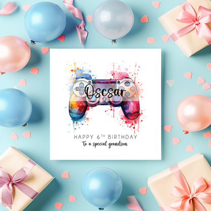 A square birthday card with a colourful watercolour game controller and the name 'Oscar' in script, surrounded by pastel balloons, gift boxes, and confetti on a light blue background.