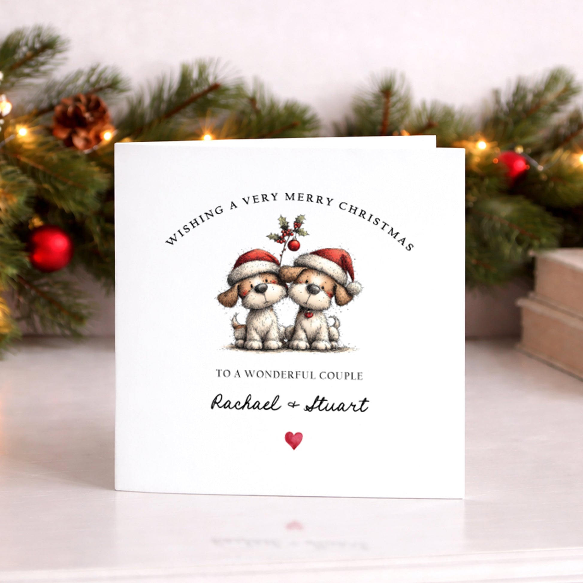 A square white Christmas card featuring an illustration of two fluffy dogs wearing red Santa hats, sitting side by side beneath a holly sprig with red berries. The text "Wishing a Very Merry Christmas" curves around the top of the illustration in spaced capitals, with "to a Special Brother & Sister-in-Law" below in smaller spaced capitals, followed by "Rachael & Stuart" in flowing cursive script and a small red heart. The card is displayed upright on a white surface in front of a lit pine garland decorated 