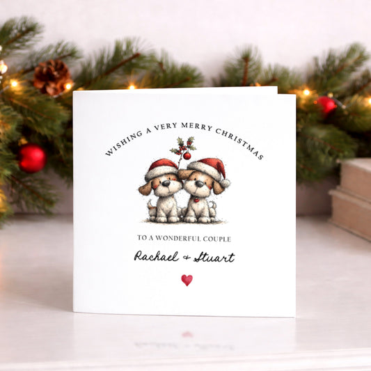 A square white Christmas card featuring an illustration of two fluffy dogs wearing red Santa hats, sitting side by side beneath a holly sprig with red berries. The text "Wishing a Very Merry Christmas" curves around the top of the illustration in spaced capitals, with "to a Special Brother & Sister-in-Law" below in smaller spaced capitals, followed by "Rachael & Stuart" in flowing cursive script and a small red heart. The card is displayed upright on a white surface in front of a lit pine garland decorated 