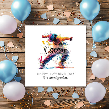 A square white birthday card featuring a vibrant watercolour illustration of a male street dancer mid-move, arms outstretched and legs wide apart, wearing an orange hoodie, blue cap, baggy jeans and trainers, surrounded by colourful paint splashes in yellow, pink, blue and red. The name "Oscar" is overlaid in bold cursive script across the illustration. Below, the text reads "Happy 12th Birthday" in spaced capitals, followed by "to a special grandson" in flowing script. The card is displayed on a wooden sur