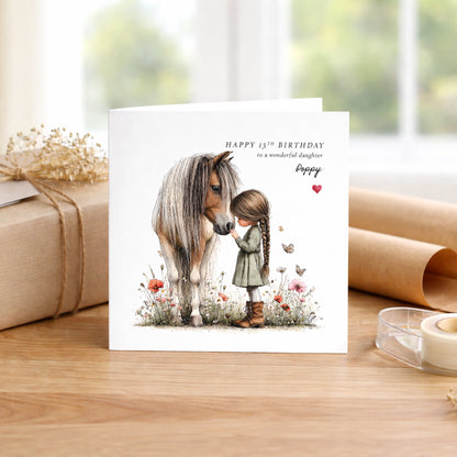 A square white birthday card featuring a watercolour illustration of a young girl with a long brown plait in a green dress and boots, tenderly touching noses with a palomino pony in a wildflower meadow with red poppies and butterflies. Text reads "Happy 13th Birthday to a wonderful daughter" with "Poppy" in cursive script and a small red heart. Displayed on a wooden surface with kraft wrapping and dried flowers.