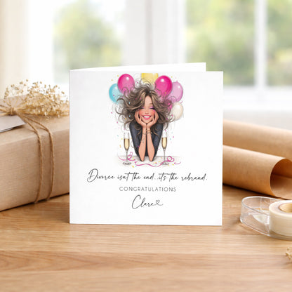 A square white greetings card featuring an illustration of a smiling woman with voluminous hair, resting her chin on her hands, surrounded by colourful pink, blue, yellow and white balloons and two champagne flutes with confetti scattered around her. The card reads "Divorce isn't the end...it's the rebrand." in cursive script, followed by "Congratulations" in spaced capitals and the name "Clare" with a small heart beneath. The card is displayed upright on a warm wooden surface alongside a kraft wrapped gift