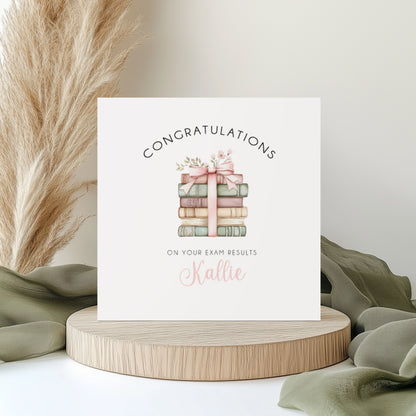 A square white card featuring a watercolour illustration of a stack of books in soft sage green, blush pink and cream, tied with a pink satin ribbon and bow with small floral details on top. Text reads "Congratulations" in arched spaced capitals above the illustration and "on Your Exam Results" below, with the name "Kallie" in pink cursive script. Displayed on a wooden disc with sage green linen and pampas grass.