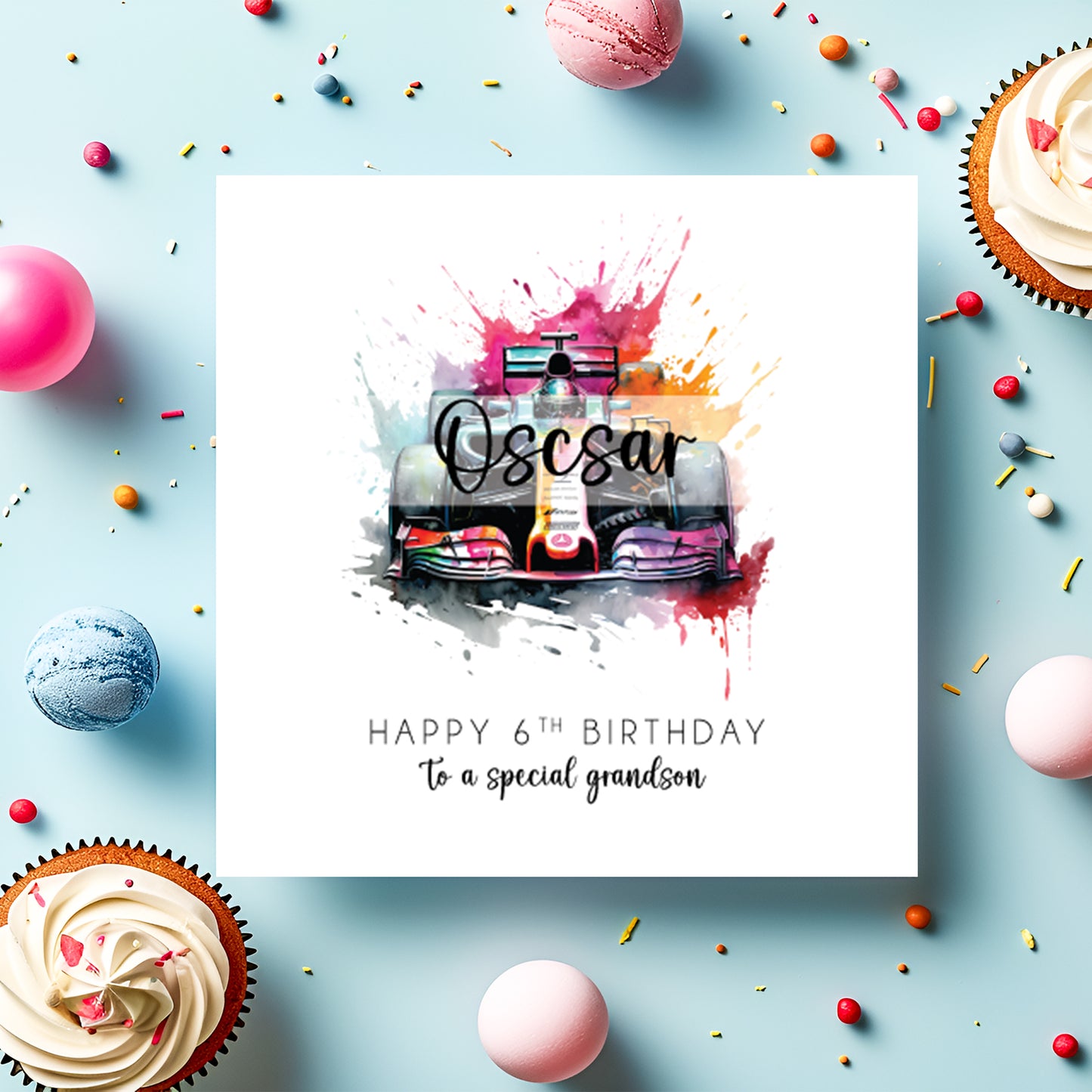A square white birthday card featuring a vibrant watercolour illustration of a Formula 1 racing car viewed head-on, surrounded by bold paint splashes in pink, purple, orange and teal. The name "Oscar" is overlaid in bold cursive script across the car. Text reads "Happy 6th Birthday to a special grandson" in spaced capitals and flowing script below. Displayed on a light blue surface with cupcakes, macarons and colourful sprinkles.