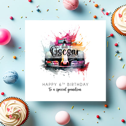 A square white birthday card featuring a vibrant watercolour illustration of a Formula 1 racing car viewed head-on, surrounded by bold paint splashes in pink, purple, orange and teal. The name "Oscar" is overlaid in bold cursive script across the car. Text reads "Happy 6th Birthday to a special grandson" in spaced capitals and flowing script below. Displayed on a light blue surface with cupcakes, macarons and colourful sprinkles.