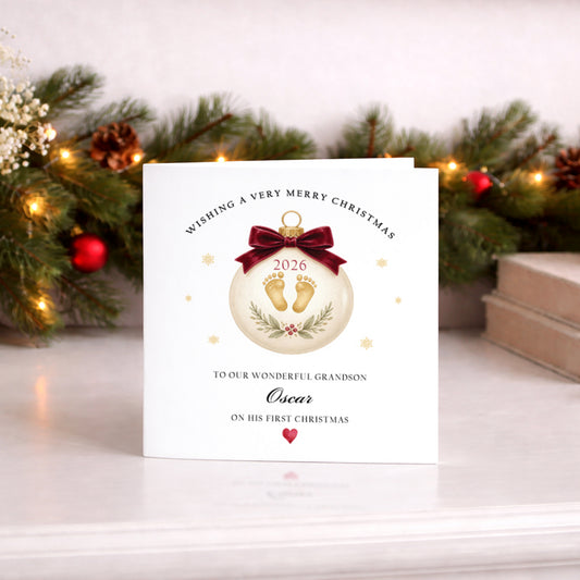 Personalised baby's first Christmas card featuring an elegant cream Christmas bauble decorated with golden baby footprints, a burgundy velvet-style bow, festive greenery and gold snowflakes. The bauble displays the year 2026, with personalised wording reading To Our Wonderful Grandson Oscar On His First Christmas and a small red heart. Suitable for baby boys and girls, with editable name, relationship and year. Supplied with a red envelope