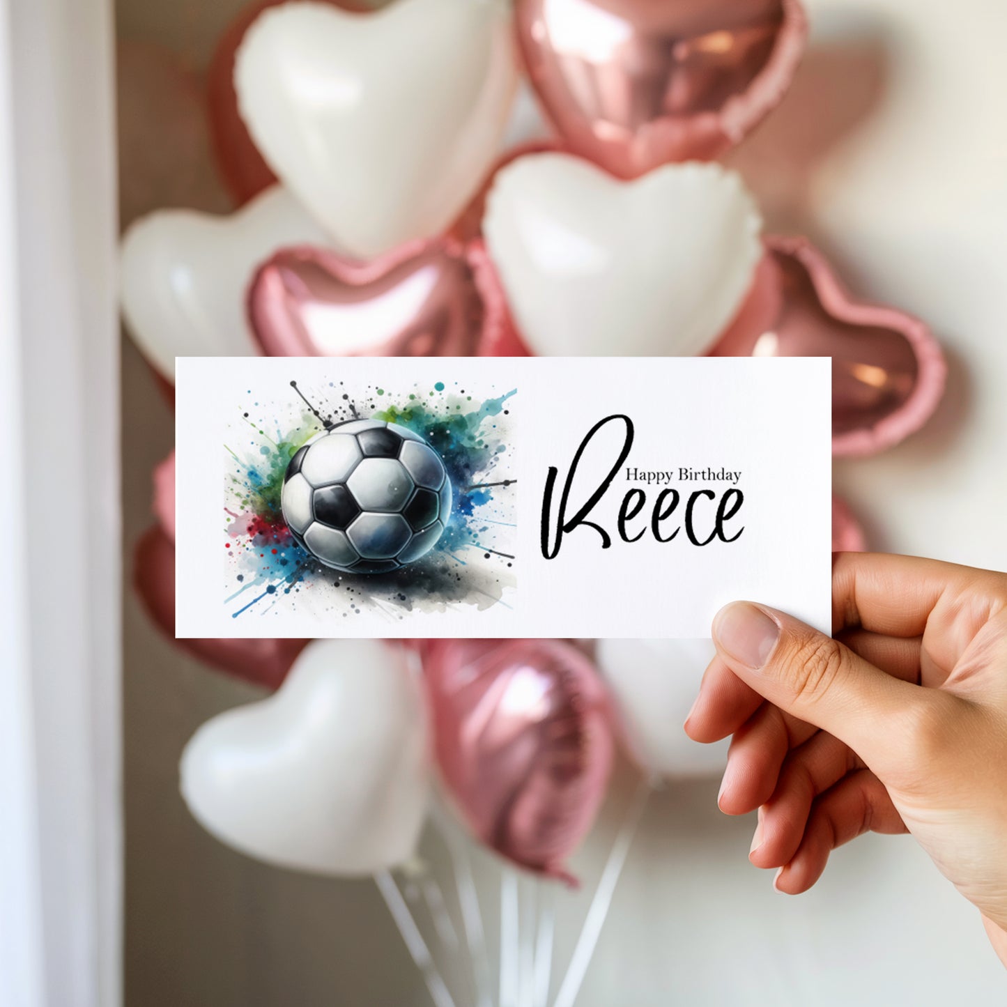 Hand holding a birthday money wallet with a football design and 'Happy Birthday (name)' text, with balloons in the background.
