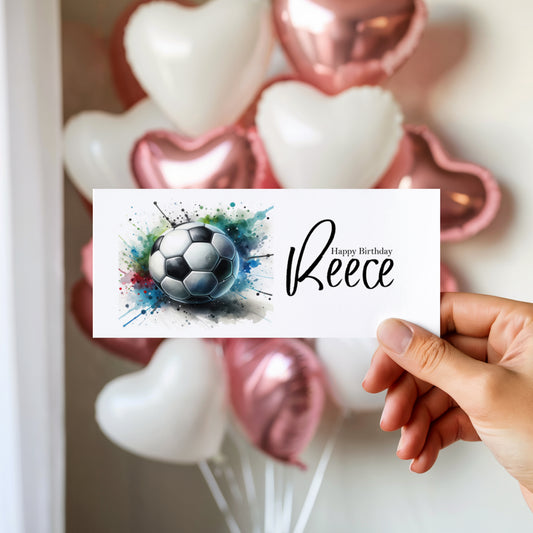 Hand holding a birthday money wallet with a football design and 'Happy Birthday (name)' text, with balloons in the background.