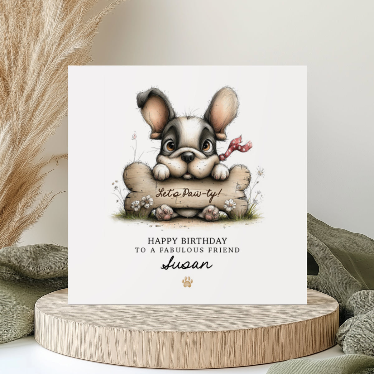 A square white birthday card featuring an illustration of a cute French Bulldog with big ears and a red polka dot bow, peering over a large bone with "Let's Paw-ty!" written on it, surrounded by small wildflowers. Text reads "Happy Birthday to a Fabulous Friend" with "Susan" in cursive script and a small gold paw print. Displayed on a wooden disc with sage green linen and pampas grass.