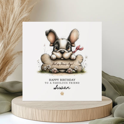A square white birthday card featuring an illustration of a cute French Bulldog with big ears and a red polka dot bow, peering over a large bone with "Let's Paw-ty!" written on it, surrounded by small wildflowers. Text reads "Happy Birthday to a Fabulous Friend" with "Susan" in cursive script and a small gold paw print. Displayed on a wooden disc with sage green linen and pampas grass.