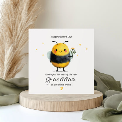 Cute Father’s Day greeting card featuring a smiling watercolour-style bumblebee holding flowers, surrounded by small heart details. The card reads “Happy Father’s Day, Thank you for bee-ing the best Grandad in the whole world.” Displayed on a neutral styled background with pampas grass and soft fabric accents.