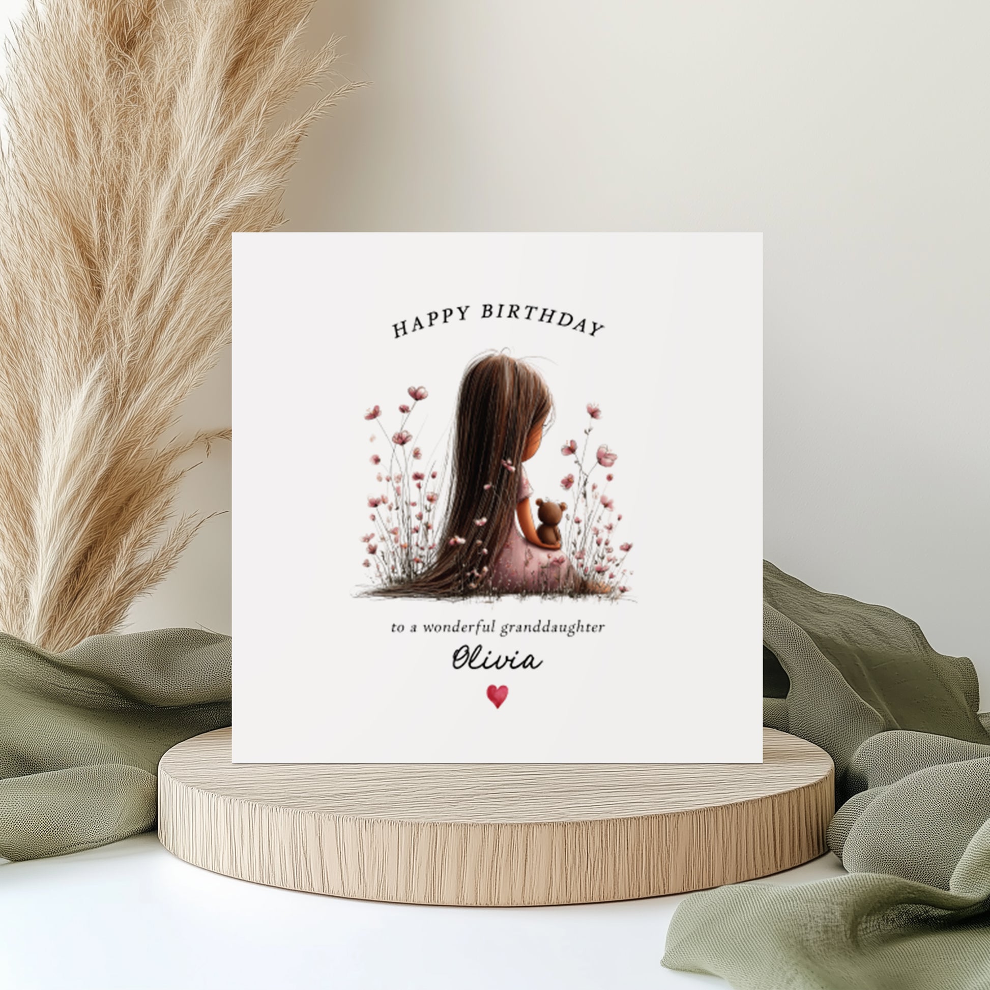 A square white birthday card featuring an illustration of a young girl with long dark hair viewed from behind, sitting in a pink wildflower meadow hugging a small teddy bear. Text reads "Happy Birthday to a wonderful granddaughter" with "Olivia" in cursive script and a small red heart. Displayed on a wooden surface with kraft wrapping and dried flowers.