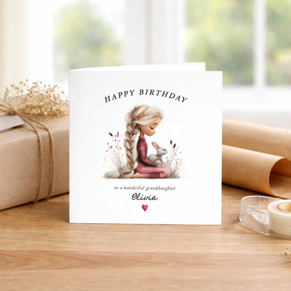 A square white birthday card featuring an illustration of a girl with a long blonde plait wearing a burgundy outfit, sitting cross-legged amongst delicate wildflowers holding a small white bunny. Text reads "Happy Birthday to a wonderful granddaughter" with "Olivia" in cursive script and a small red heart. Displayed on a wooden surface with kraft wrapping and dried flowers.