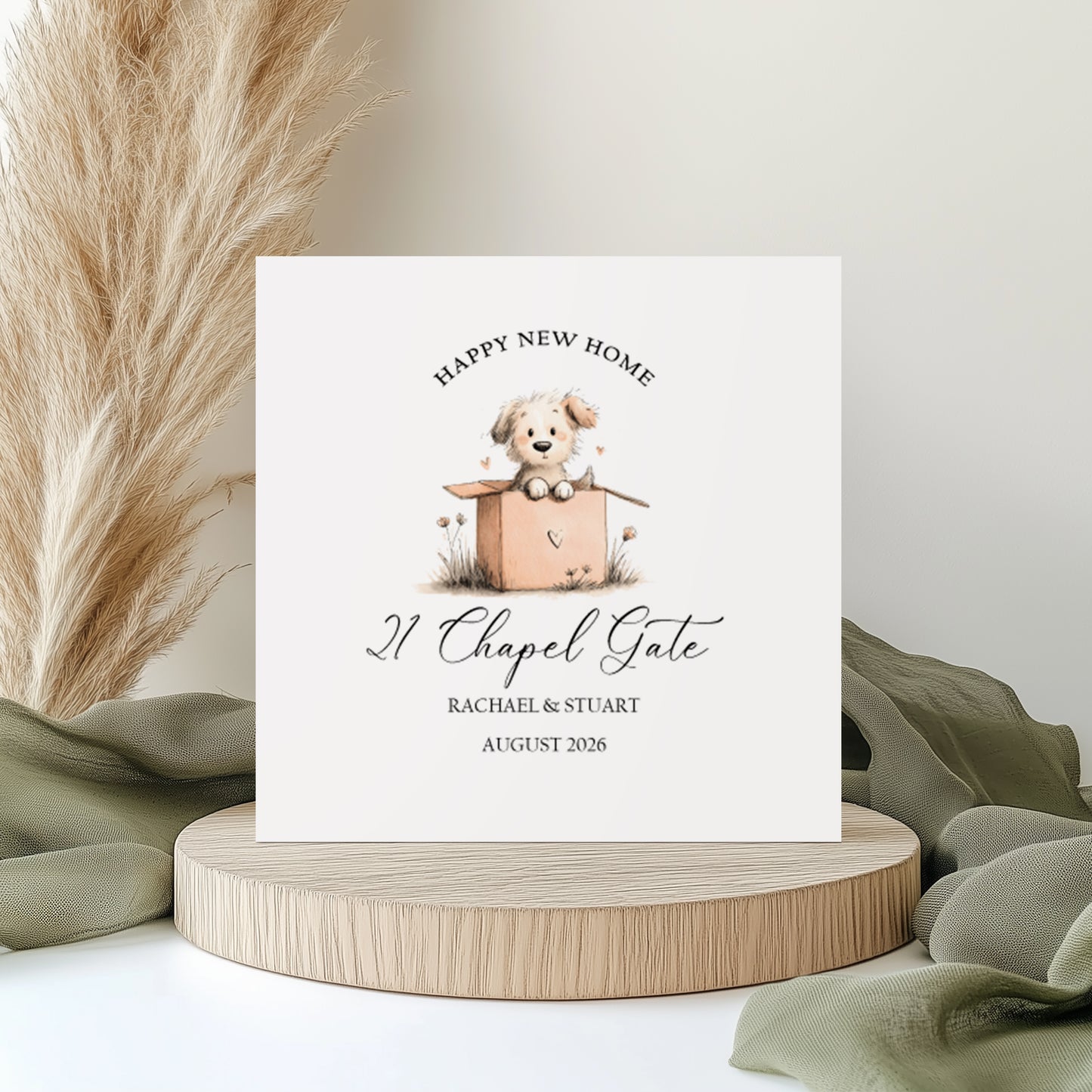 A square white new home card featuring a watercolour illustration of a puppy in a moving box. Text reads "Happy New Home" in arched capitals, with "21 Chapel Gate" in cursive script, "Rachael & Stuart" and "August 2026" below. Displayed on a wooden disc with sage green linen and pampas grass.