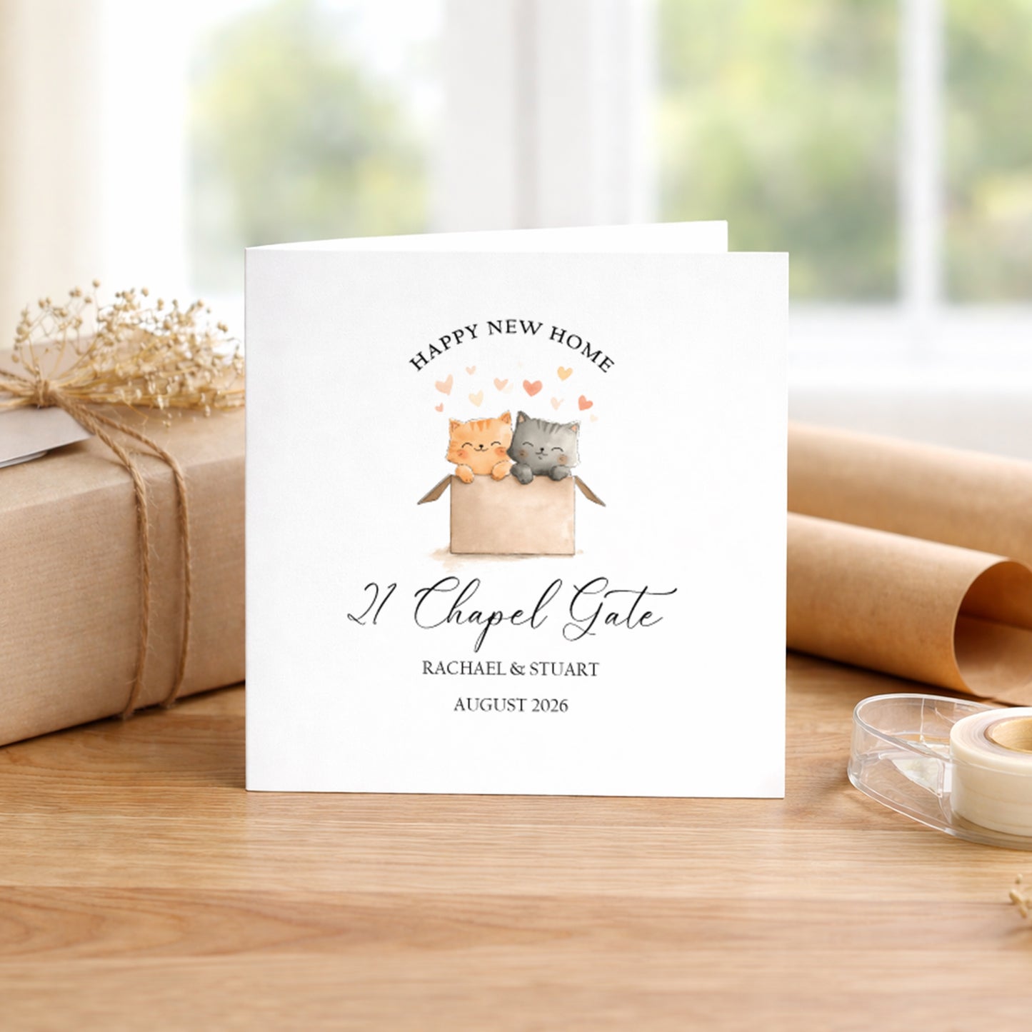 A personalised white greeting card featuring a watercolor illustration of two cats in a box with hearts and the text 'HAPPY NEW HOME 21 Chapel Gate RACHAEL & STUART AUGUST 2026', displayed on a wooden table with a wrapped gift and paper rolls in the background.