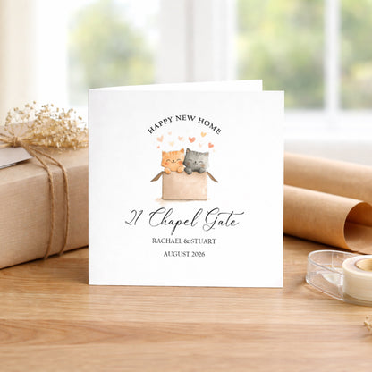 A personalised white greeting card featuring a watercolor illustration of two cats in a box with hearts and the text 'HAPPY NEW HOME 21 Chapel Gate RACHAEL & STUART AUGUST 2026', displayed on a wooden table with a wrapped gift and paper rolls in the background.