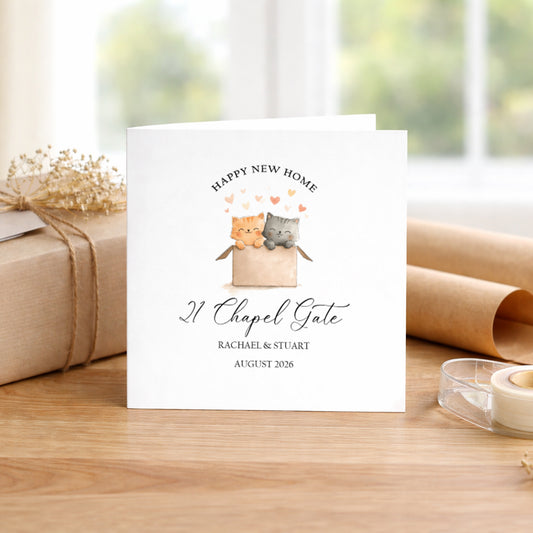 A personalised white greeting card featuring a watercolor illustration of two cats in a box with hearts and the text 'HAPPY NEW HOME 21 Chapel Gate RACHAEL & STUART AUGUST 2026', displayed on a wooden table with a wrapped gift and paper rolls in the background.