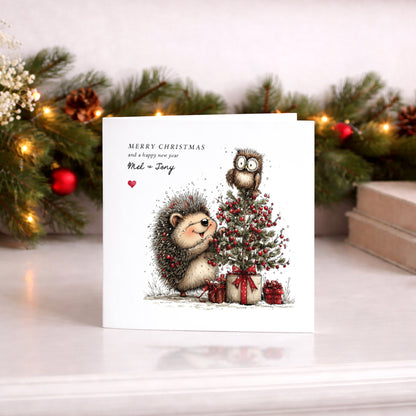 A square white Christmas card featuring an illustration of a smiling hedgehog decorating a snow-dusted Christmas tree adorned with red berries and wrapped gifts below, with a wide-eyed owl perched on top. Text reads "Merry Christmas and a happy new year" with "Mel & Tony" in cursive script and a small red heart. Displayed against a lit pine garland with red baubles.