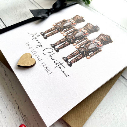 A white Christmas greeting card featuring three illustrated nutcracker figures and the text 'Merry Christmas to a special family,' styled on a light wood surface with a black ribbon and a small gold heart.