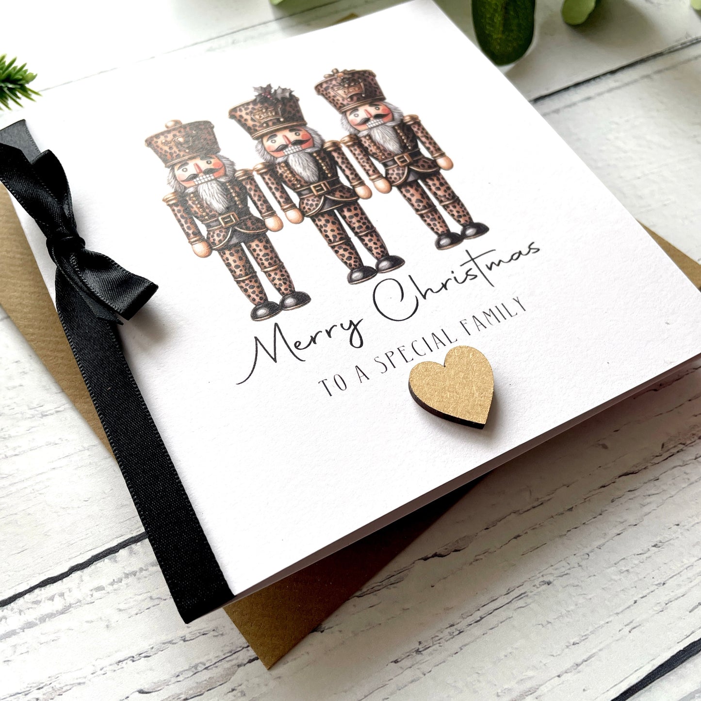 A white Christmas greeting card featuring three illustrated nutcracker figures, the text 'Merry Christmas to a special family,' a black ribbon bow, and a small gold heart, photographed on a white wood surface with a kraft envelope partially visible underneath.