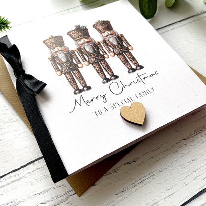 A white Christmas greeting card featuring three illustrated nutcracker figures, the text 'Merry Christmas to a special family,' a black ribbon bow, and a small gold heart, photographed on a white wood surface with a kraft envelope partially visible underneath.