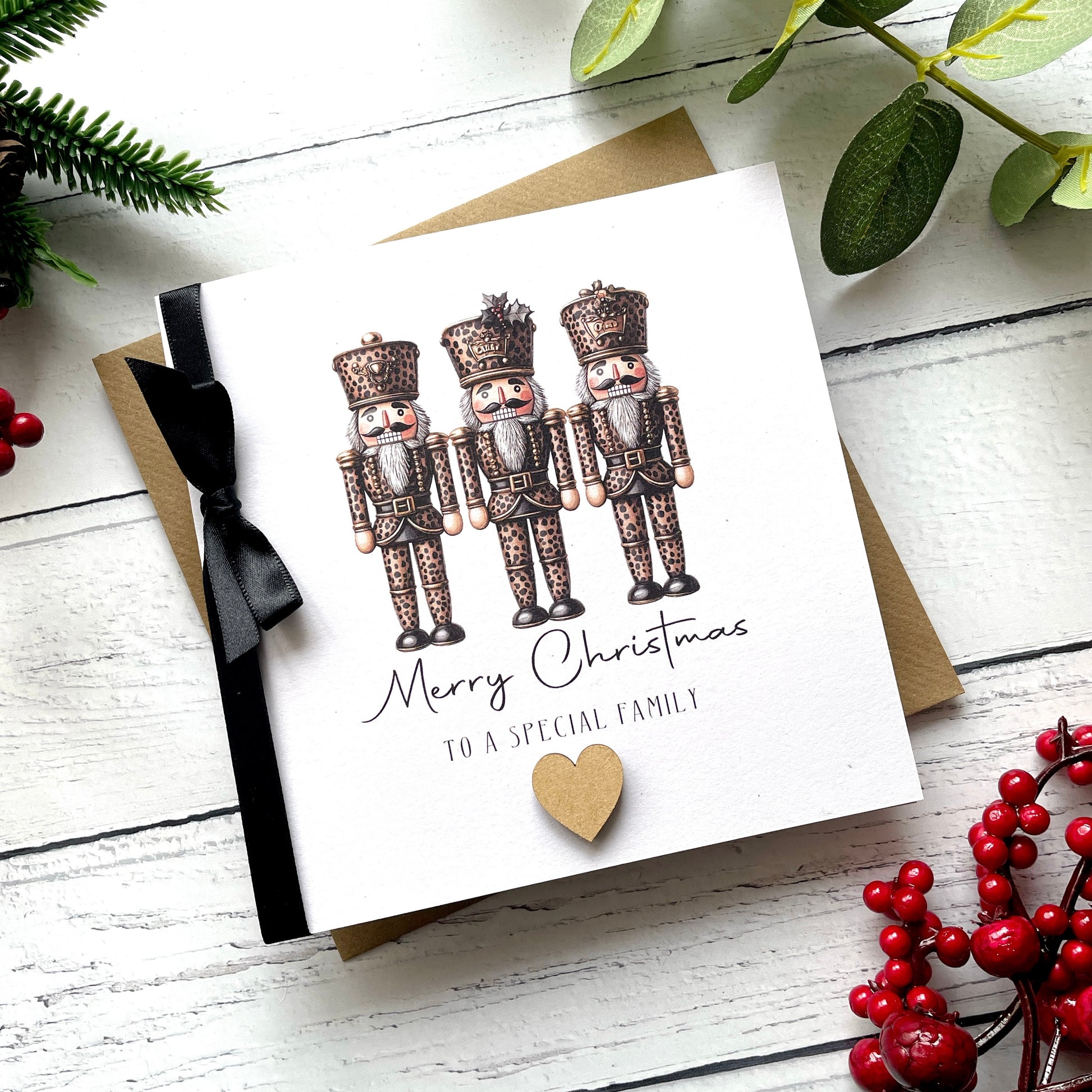 A square Christmas greeting card with an illustration of four nutcrackers, the text 'Merry Christmas to a special family,' a black ribbon, and a small gold heart, photographed on a white wood surface with a kraft envelope behind it and holiday greenery and red berries around it.