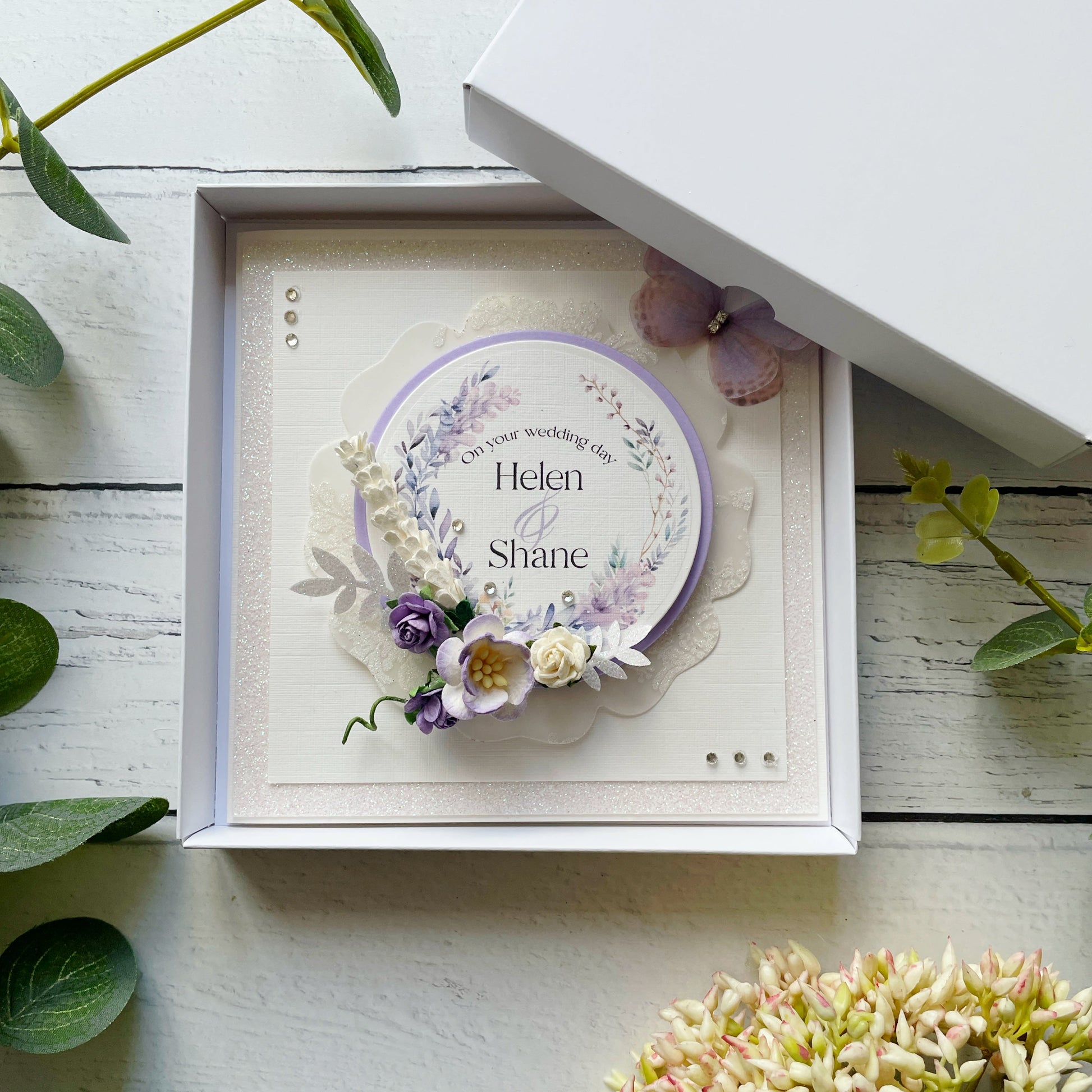 Decorative wedding card with lilac floral elements in a box, personalised text details, on a wooden surface with flowers around.