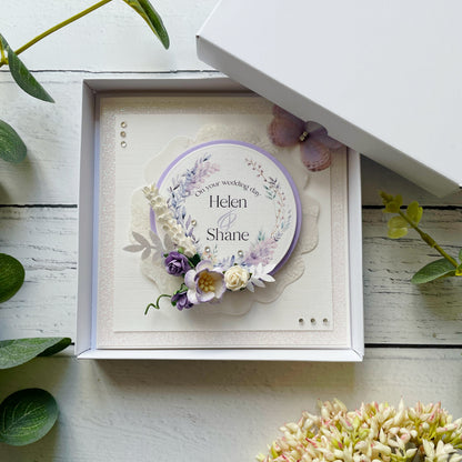 Decorative wedding card with lilac floral elements in a box, personalised text details, on a wooden surface with flowers around.