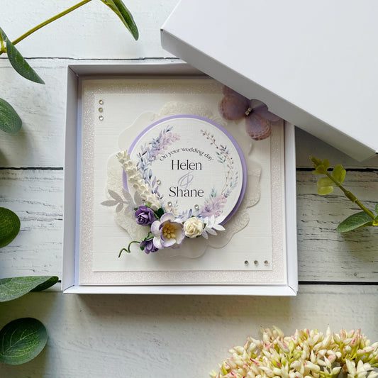 Decorative wedding card with lilac floral elements in a box, personalised text details, on a wooden surface with flowers around.