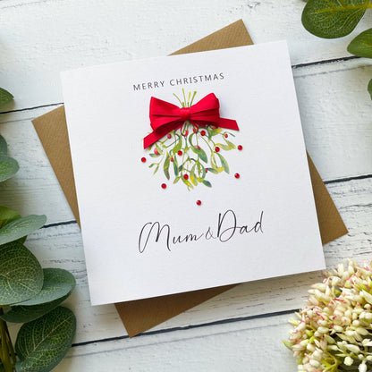 A square white Christmas greeting card with 'MERRY CHRISTMAS' above a watercolor mistletoe design with a red bow, and 'Mum & Dad' in script below, shown on top of a brown envelope on a white wood surface with green leaves and flowers around it.