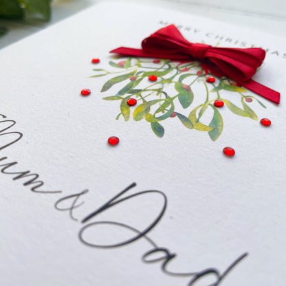 A close-up of a white greeting card with elegant black script reading 'Mum & Dad,' decorated with a watercolor mistletoe design in green and red, and a red ribbon bow on top.